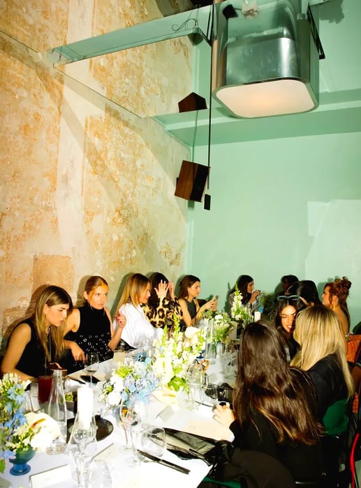 Chic women gather around a long dining table beneath a mirrored ceiling.