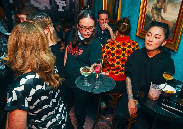 A person with shoulder-length hair and glasses carries a tray of drinks in a bustling bar.