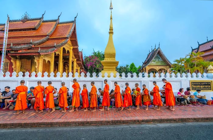 Monks dressed in orange robes line up to receive alms in front of a golden stupa on the streets of Luang Prabang, Laos.