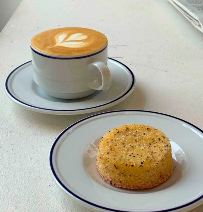 A latte adorned with leaf-shaped foam beside a small cake dusted with sugar.