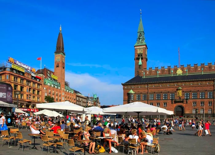 People gather at café tables shaded by large white umbrellas in a bustling city square surrounded by buildings.