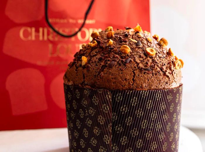 A rich dark panettone garnished with chopped nuts, next to a branded bakery bag.