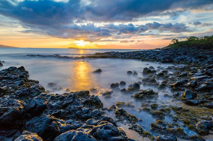 Waves crash against the volcanic rocks as the sun sets over the horizon.