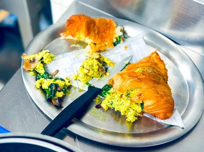 An egg-laden croissant sandwich in a somewhat messy state on a silver platter.