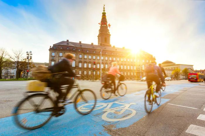 Cyclists blur past on a bike lane in Copenhagen as the sun sets over Christiansborg Palace.