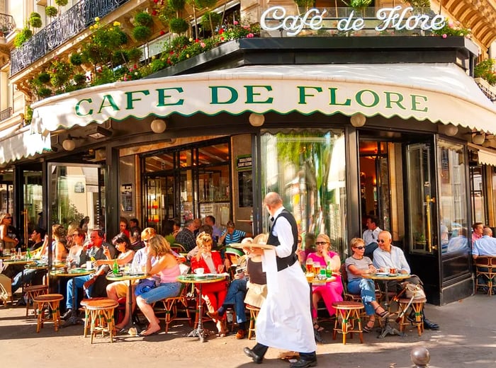 A waiter navigates in front of customers seated at outdoor tables under an awning reading Café de Flore.