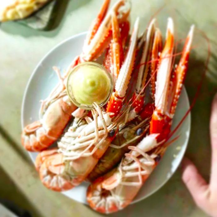 A detailed image featuring several large cooked langoustines on a plate, accompanied by a small dish of butter, resting next to the photographer's hand.