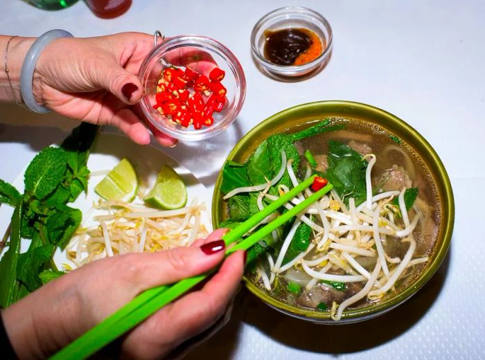 Hands sprinkle chiles into a bowl of pho using vibrant green chopsticks.