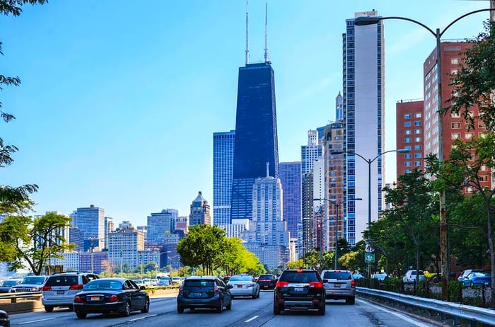 A street-level view of traffic flowing along Lake Shore Drive in Downtown Chicago, framed by skyscrapers, including the iconic Sears Tower.