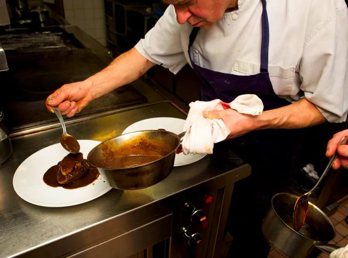 A chef drizzles sauce over a steak on a preparation table in the kitchen.