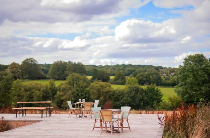 Outdoor tables arranged on a long wooden deck, set against a backdrop of rolling hills and trees on a cloudy day.