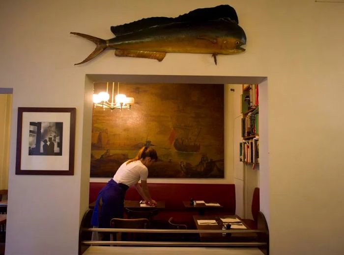 A server is seen preparing tables through a cutout in the wall, beneath a stuffed fish.