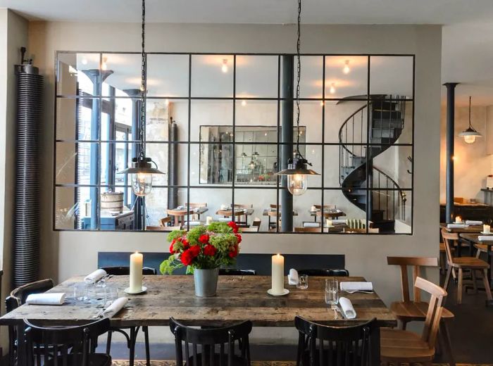 An interior shot of the restaurant showcasing rustic wooden farmhouse tables, simple seating, expansive windows dividing dining areas, and a metal spiral staircase.