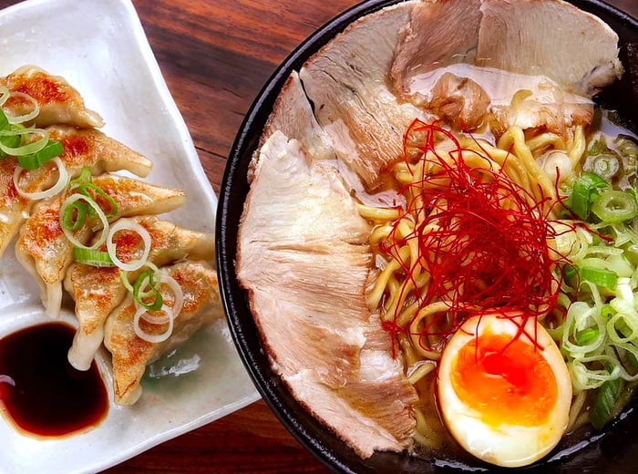 A bowl of ramen adorned with slices of pork, egg, and scallions, accompanied by a plate of gyoza with dipping sauce.