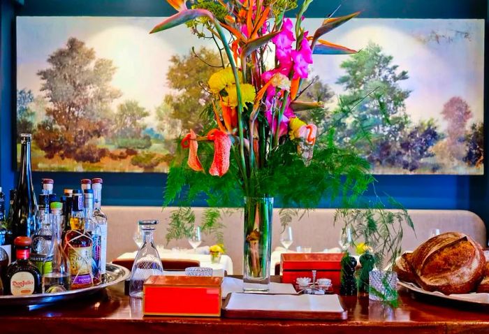 A dining room side table adorned with a vase of flowers, a basket of bread, and a selection of spirits.