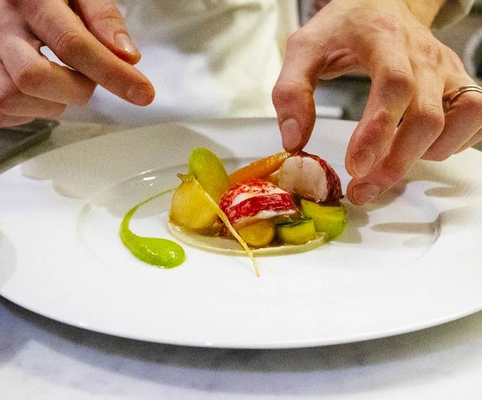 A chef arranges thick slices of lobster atop a variety of other ingredients on a mostly pristine white plate.