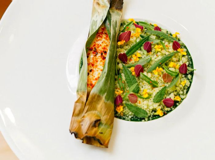 A cylinder of roasted fish wrapped in banana leaves sits beside a couscous salad adorned with fresh herbs and edible flowers.