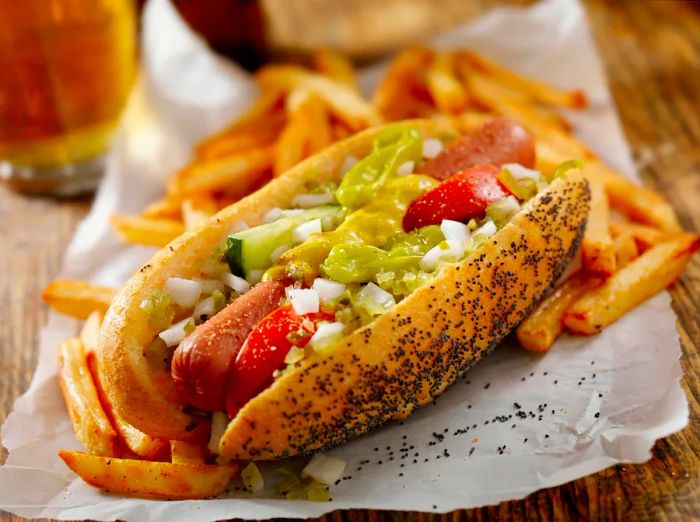 A classic Chicago hot dog served alongside fries and a beer. This hot dog features fresh tomatoes, diced onions, green relish, peppers, pickles, yellow mustard, and a sprinkle of celery salt.