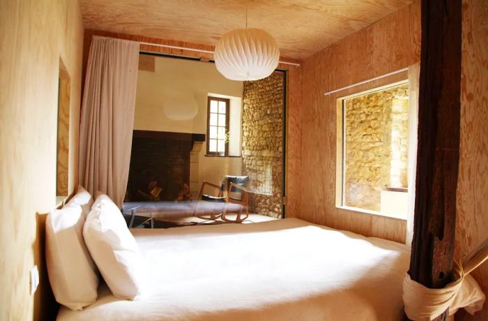 A cozy inn room featuring exposed stone walls, a freshly made bed, a decorative paper lantern, and a sizable fireplace.