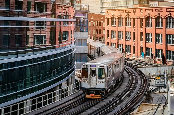 A silver commuter train glides along an elevated track amidst the buildings.