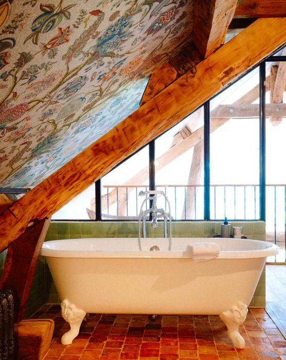 A standalone bathtub beneath a sloped ceiling adorned with illustrated wallpaper, leading to a bright patio outside.