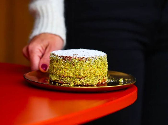 A hand sets down a small cylindrical cake adorned with finely chopped pistachios.