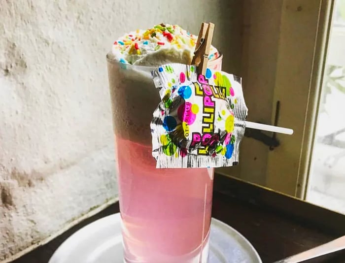 A vibrant pink cocktail served in a highball glass, adorned with whipped cream and sprinkles, featuring a lollipop clipped to the rim, resting on a small plate alongside a long ice cream spoon, set against a bar corner with a cement wall.