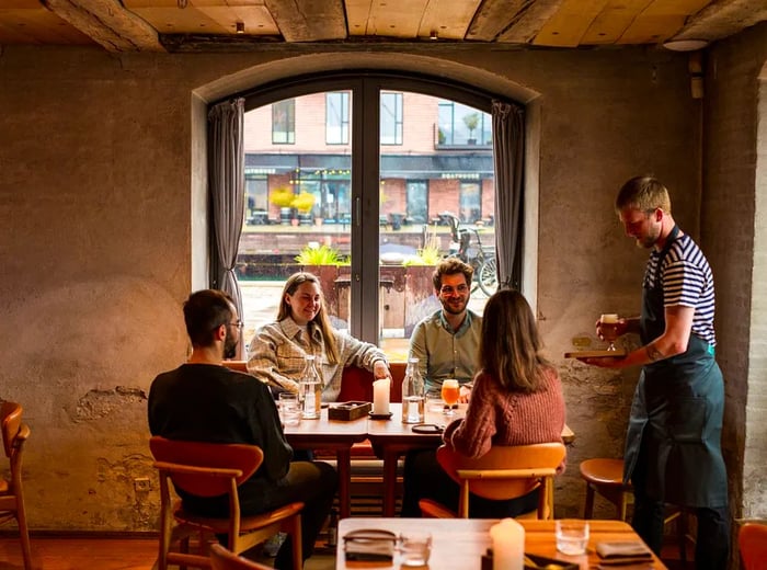 Guests enjoy their meals at a table by the window as a server brings over a drink.