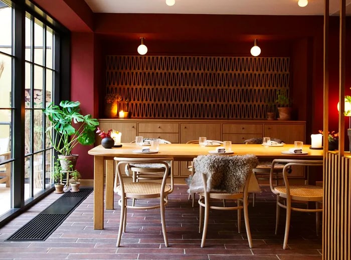 A dining room featuring a patterned wall, a credenza, wooden dividers, and a large table surrounded by slender wooden chairs, one adorned with a fur throw.