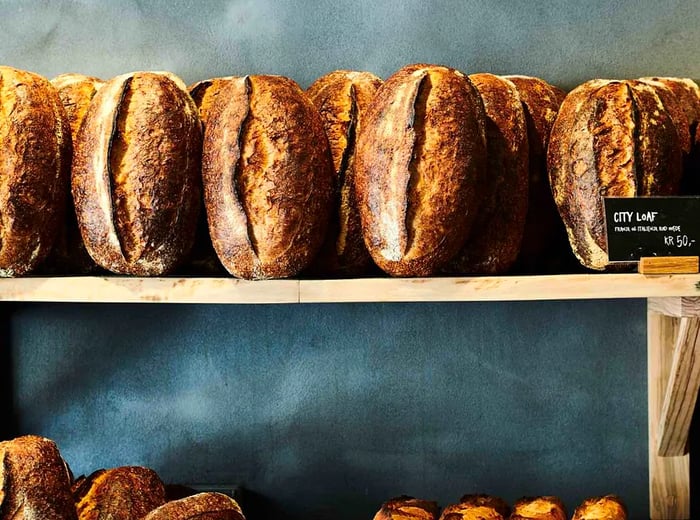 A wooden shelf displays loaves of sourdough bread leaning against a slate wall, accompanied by a small chalkboard sign that reads 'City Loaf.'