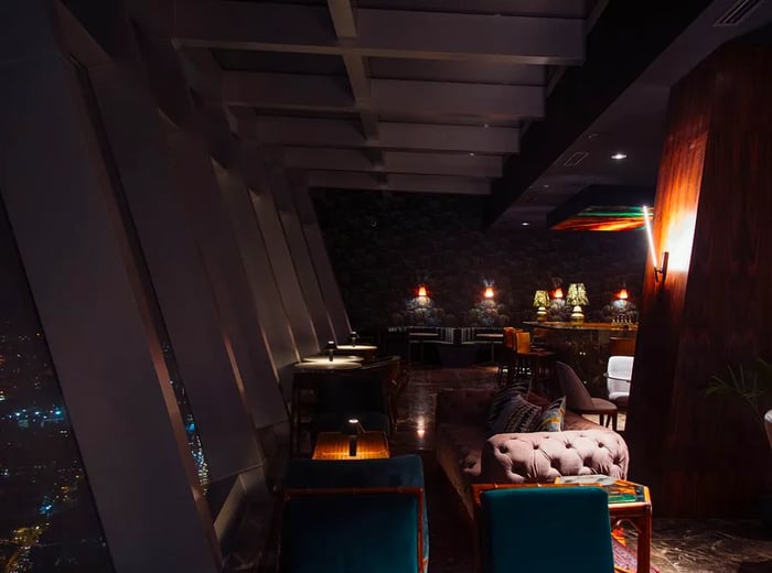 A nighttime view of the bar's interior, featuring cozy lounge seating under soft lighting and a series of angled windows that open to the cityscape below.