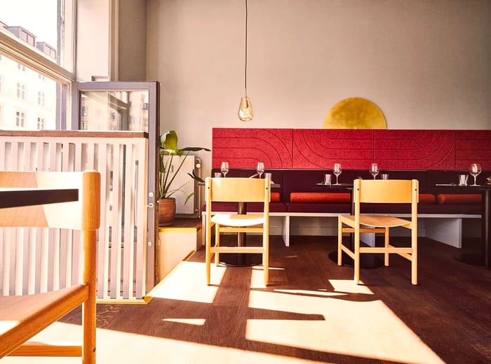 An open and airy restaurant space featuring light wood chairs arranged around tables along a tall red banquette.