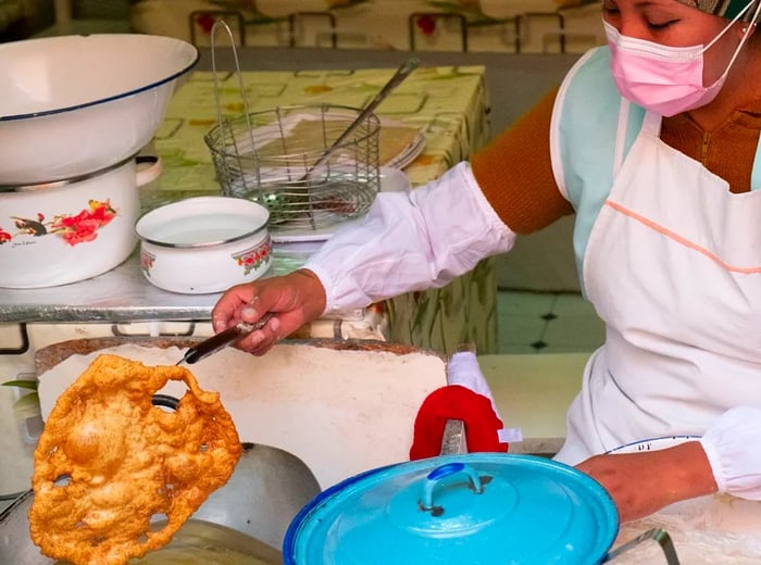 A chef uses a long fork to lift a fried buñuelo from a vibrant blue pot.