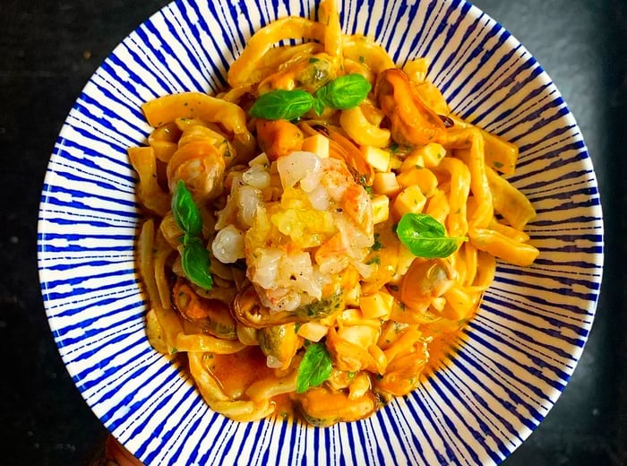 A hand holding a geometric plate filled with scialatielli pasta, generously topped with a heap of chopped seafood and scattered herbs, captured from above.