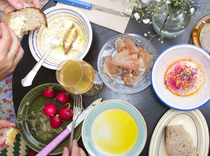 From above, two people dine at a table filled with an assortment of dishes, including whole radishes, bread, olive oil, sliced meats, alongside glasses of wine and a floral arrangement.