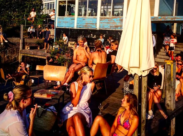 A group of people enjoys drinks on a dock outside the restaurant, with some sunbathing nearby.