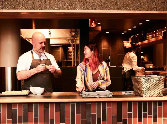 A husband and wife engage in conversation while preparing food and setting the scene behind the restaurant counter.