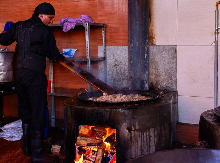 A chef skillfully stirs a massive pot of chicharrones over a wood-burning fire with a large paddle.