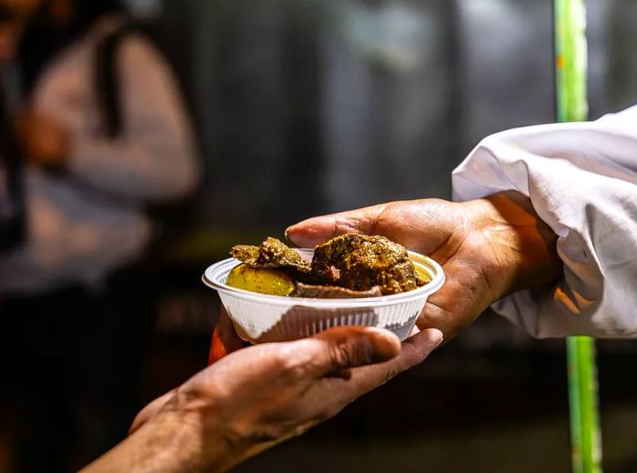 A vendor offers a small plastic bowl filled with anticuchos.