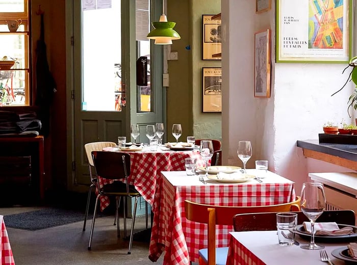 The restaurant's interior is bathed in natural light, featuring empty tables dressed in checkerboard tablecloths and set for service. The walls display a mix of art and posters, complemented by a vibrant midcentury pendant light hanging from the ceiling.