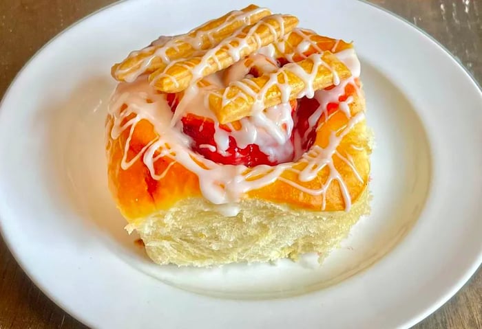 A kolache adorned with generous drizzles of icing, jam, and various toppings, presented on a white plate