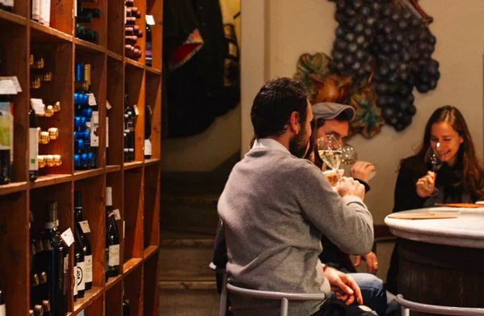 Three individuals are gathered at a table, savoring wine, with shelves of unopened bottles lining a nearby wall.