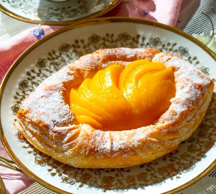 A diamond-shaped pastry filled with vibrant yellow peach slices, dusted with sugar around the edges, elegantly displayed on a decorative plate next to an ornate tea mug.