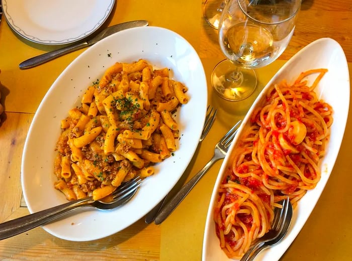 Aerial view of two pasta dishes elegantly presented on oblong plates, accompanied by a glass of wine.