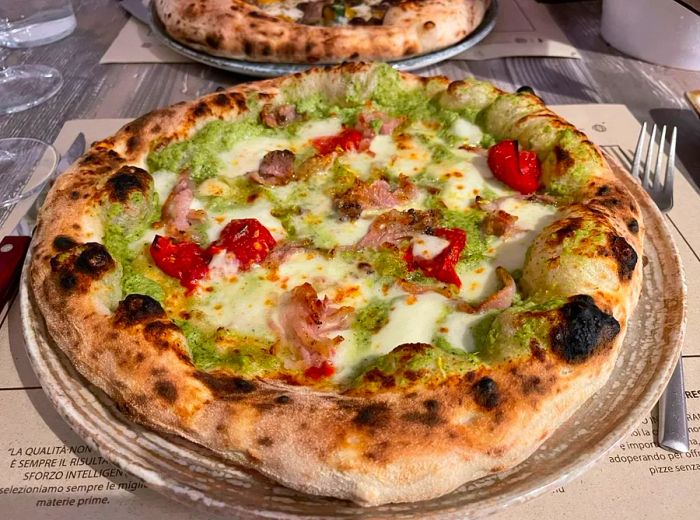 A whole pizza on a plate adorned with green sauce, cheese, blistered tomatoes, and meat