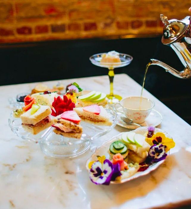 A teapot gracefully pouring tea into an ornate mug, accompanied by a platter of vibrant sandwiches and sweets, set on a marble dining table surrounded by a plush banquette.