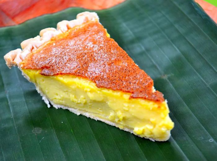 A slice of custard pie with a vibrant yellow filling and a caramelized top, elegantly served on a banana leaf.