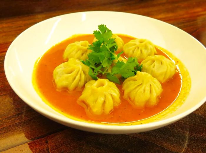 A bowl filled with momos drenched in a light orange sauce, garnished with a sprig of cilantro.