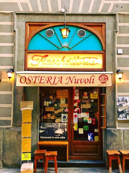 The exterior of a restaurant featuring a large awning under a vibrant art deco sign, with windows displaying various signs and a few wooden stools positioned out front.