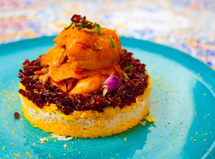 Tender chicken pieces drizzled with sauce atop a bed of rice, garnished with barberries and a rose petal on a vibrant ceramic plate.